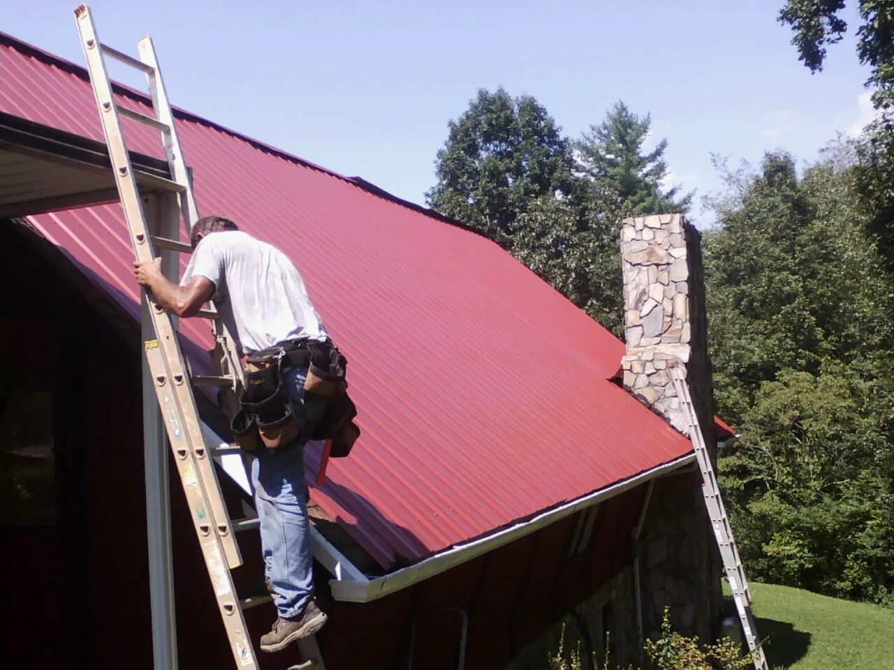 Roofer on a ladder beside a red metal roof and stone chimney — Soffit & Fascia Repair by Roofing Brothers in Maryville